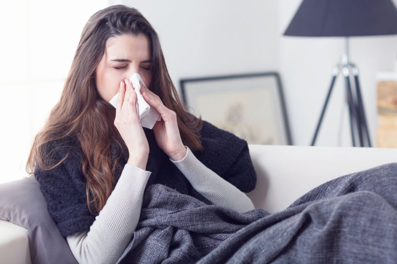 Woman sneezing on couch and realizing that her indoor air quality is affecting her allergies.