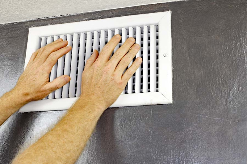 Why Is My AC Blowing Hot Air? A pair of adult male hands feeling the flow of air coming out of an air vent on a wall near a ceiling. Man with hands in front of an air vent feeling for air flow.